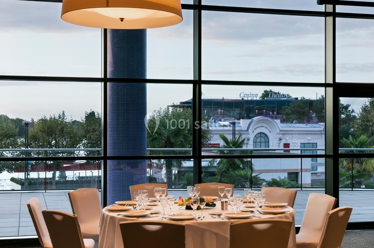 Table dressée avec vaisselle élégante dans une salle lumineuse, vue sur des palmiers et un bâtiment à travers de grandes…