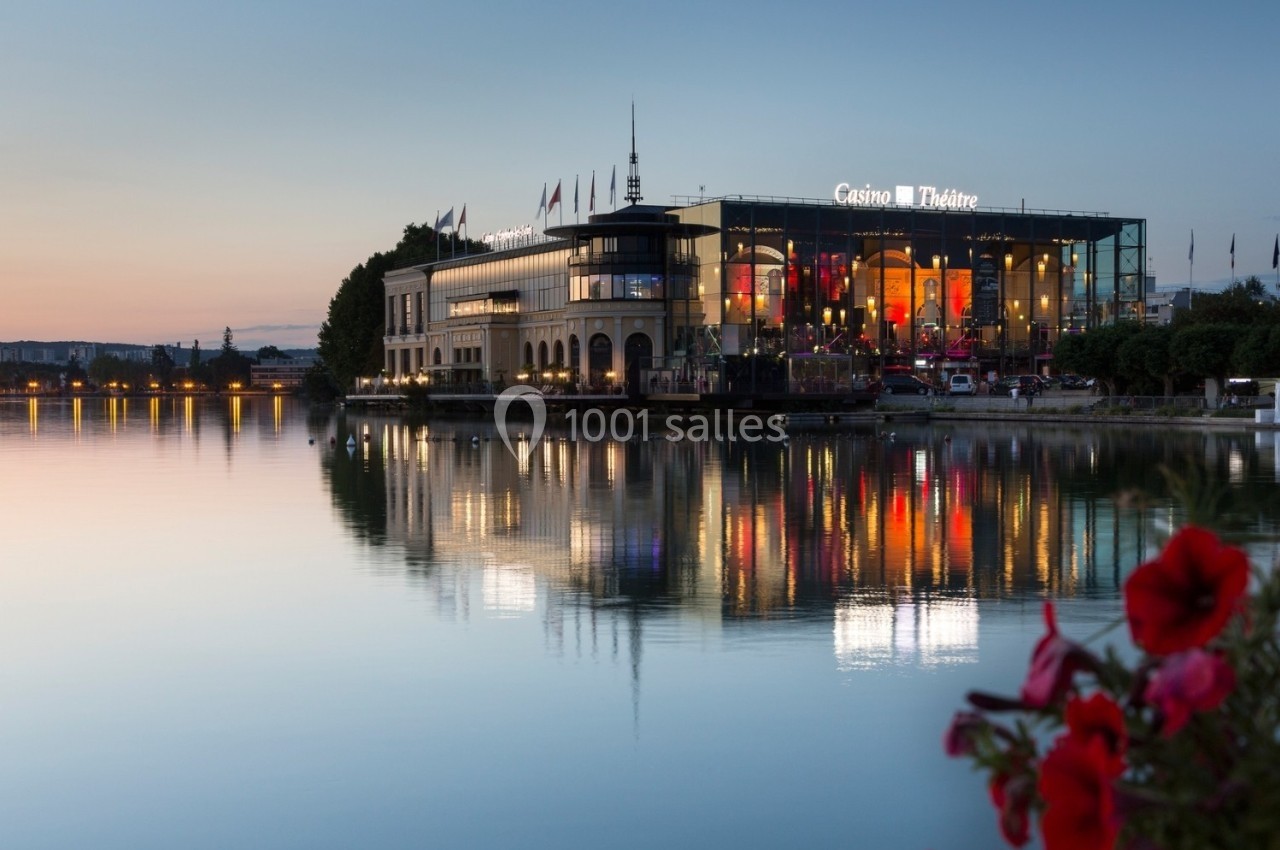 Façade illuminée d'un casino-théâtre au bord d'un lac au crépuscule, avec reflets sur l'eau et fleurs au premier plan.