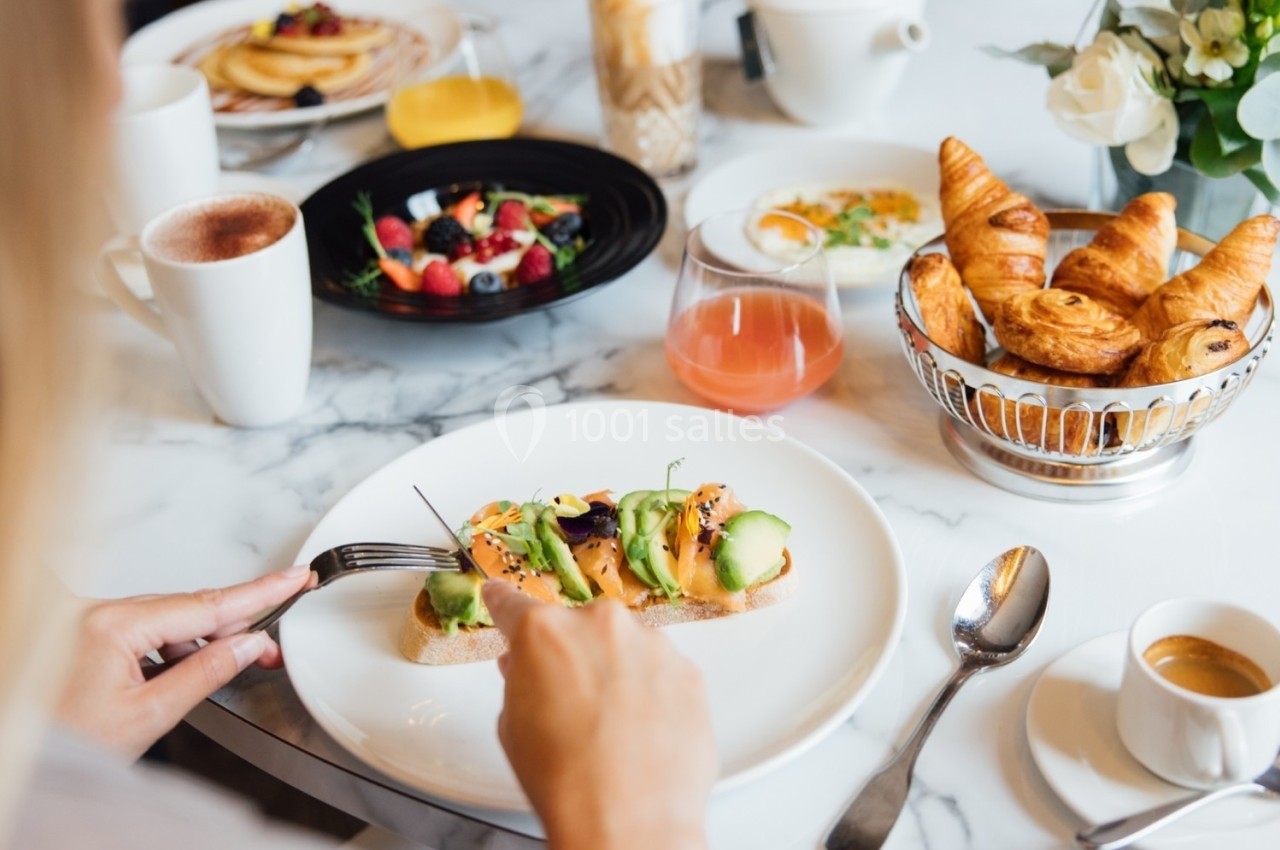 Une personne déguste un toast à l'avocat avec des croissants, fruits et boissons sur une table de petit-déjeuner.