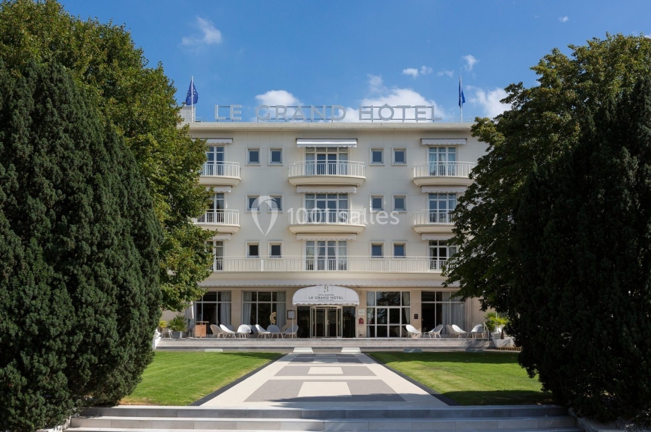 Façade d'un grand hôtel blanc entouré d'arbres, avec une entrée centrale et un chemin pavé menant à l'entrée.