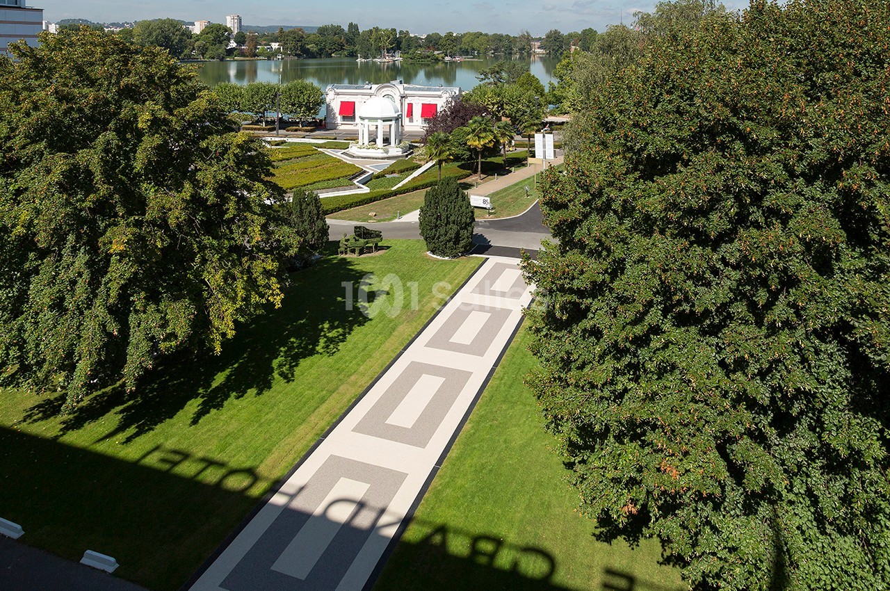 Vue aérienne d'un parc avec allées géométriques, pelouses, arbres et un bâtiment blanc près d'un plan d'eau.