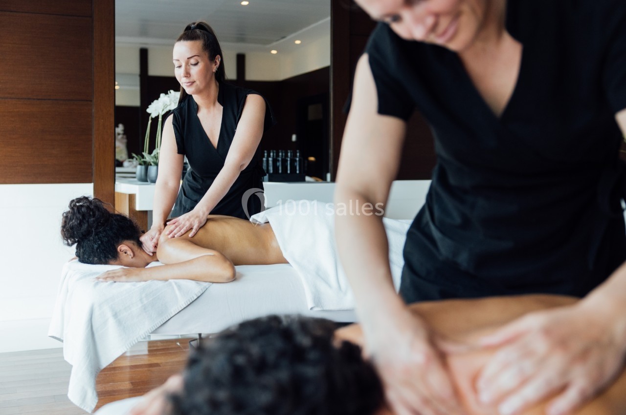 Deux praticiennes effectuent des massages sur deux personnes allongées dans une salle lumineuse au décor épuré.