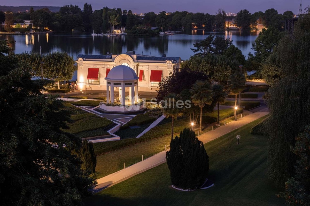 Vue nocturne d'un parc avec un pavillon éclairé, des allées bordées d'arbres et un plan d'eau en arrière-plan.