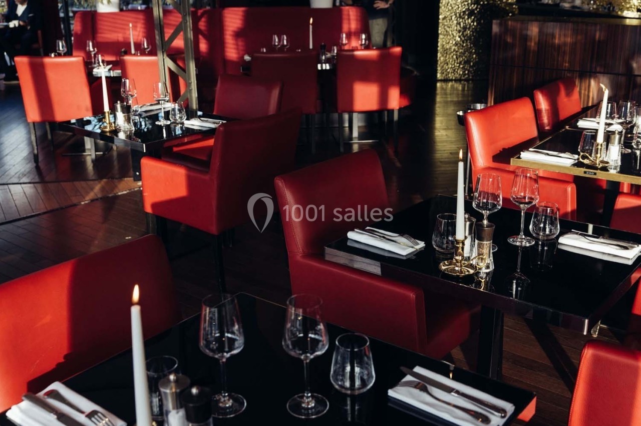 Salle de restaurant élégante avec des tables dressées, des chaises rouges et une lumière naturelle en fin de journée.