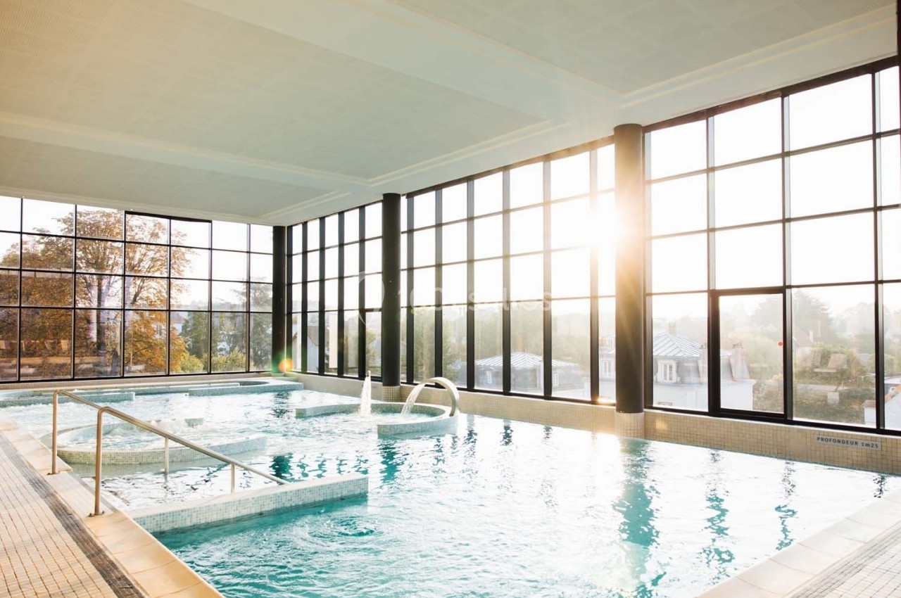 Piscine intérieure lumineuse avec de grandes baies vitrées laissant entrer la lumière du soleil, entourée de verdure.