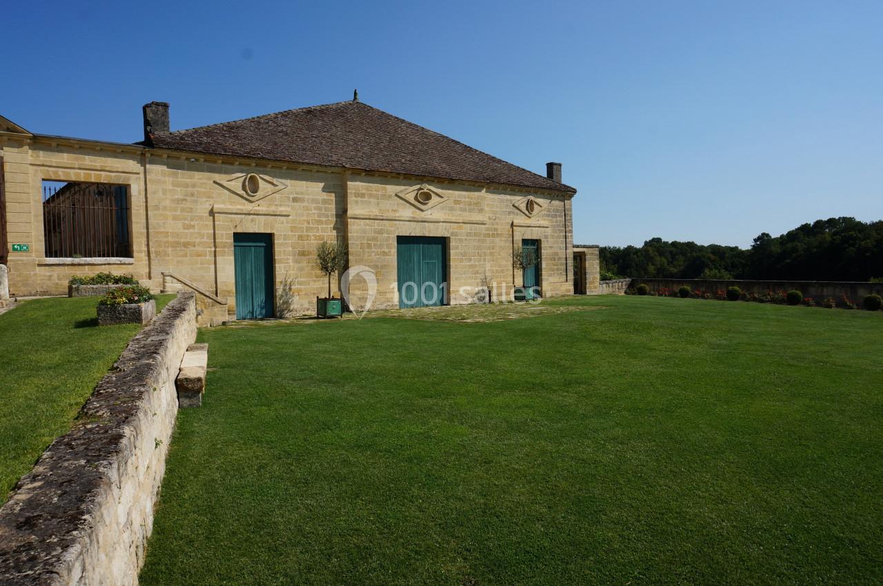 Bâtiment en pierre avec toit en pente, grandes portes vertes et pelouse bien entretenue sous un ciel bleu.