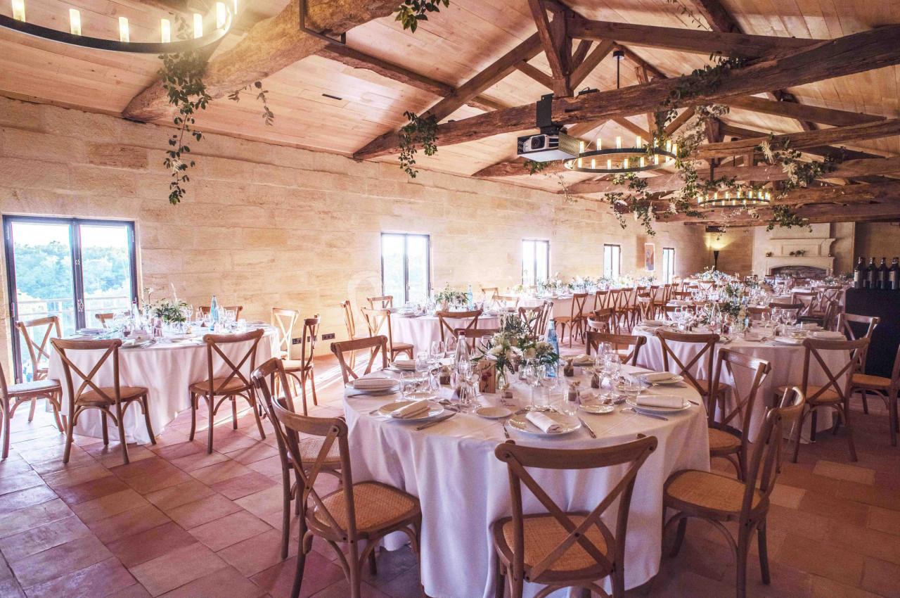 Salle de réception lumineuse avec tables rondes dressées, chaises en bois et poutres apparentes au plafond.