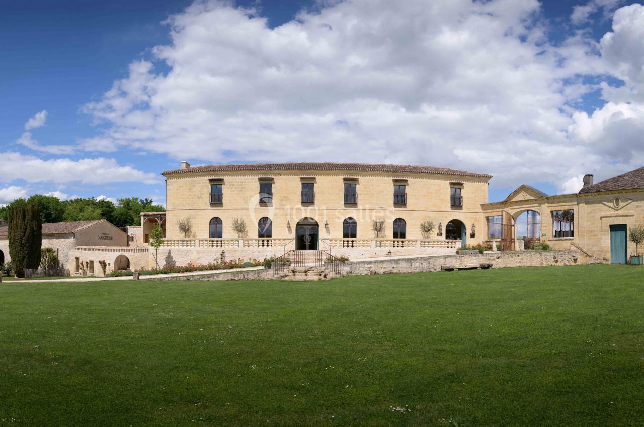 Bâtiment en pierre de style classique entouré de verdure, sous un ciel partiellement nuageux.