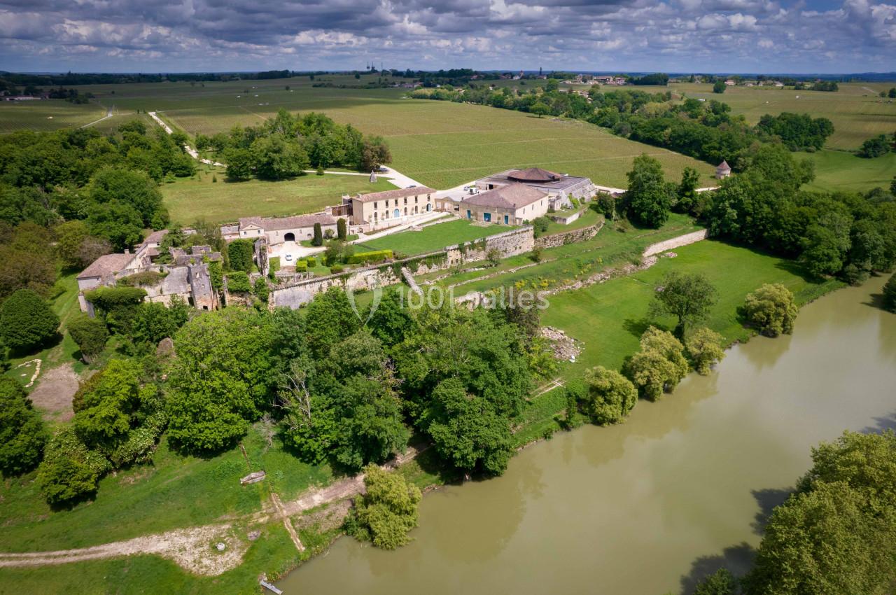 Vue aérienne d'un domaine entouré de vignes, de bâtiments en pierre et d'un cours d'eau bordé de verdure.