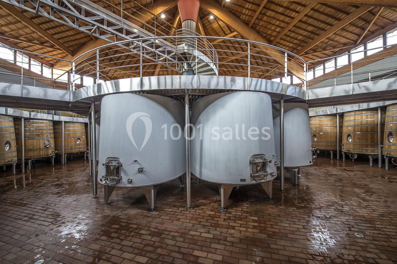 Cuves en acier inoxydable dans une cave vinicole, entourées de tonneaux en bois et situées sur un sol en briques.