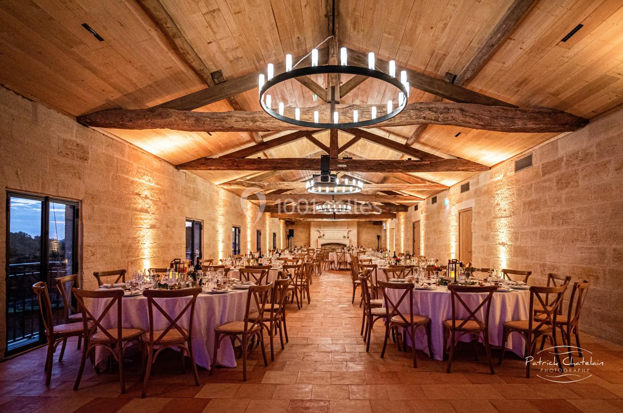 Salle de réception avec tables rondes dressées, chaises en bois, poutres apparentes et lustres suspendus.