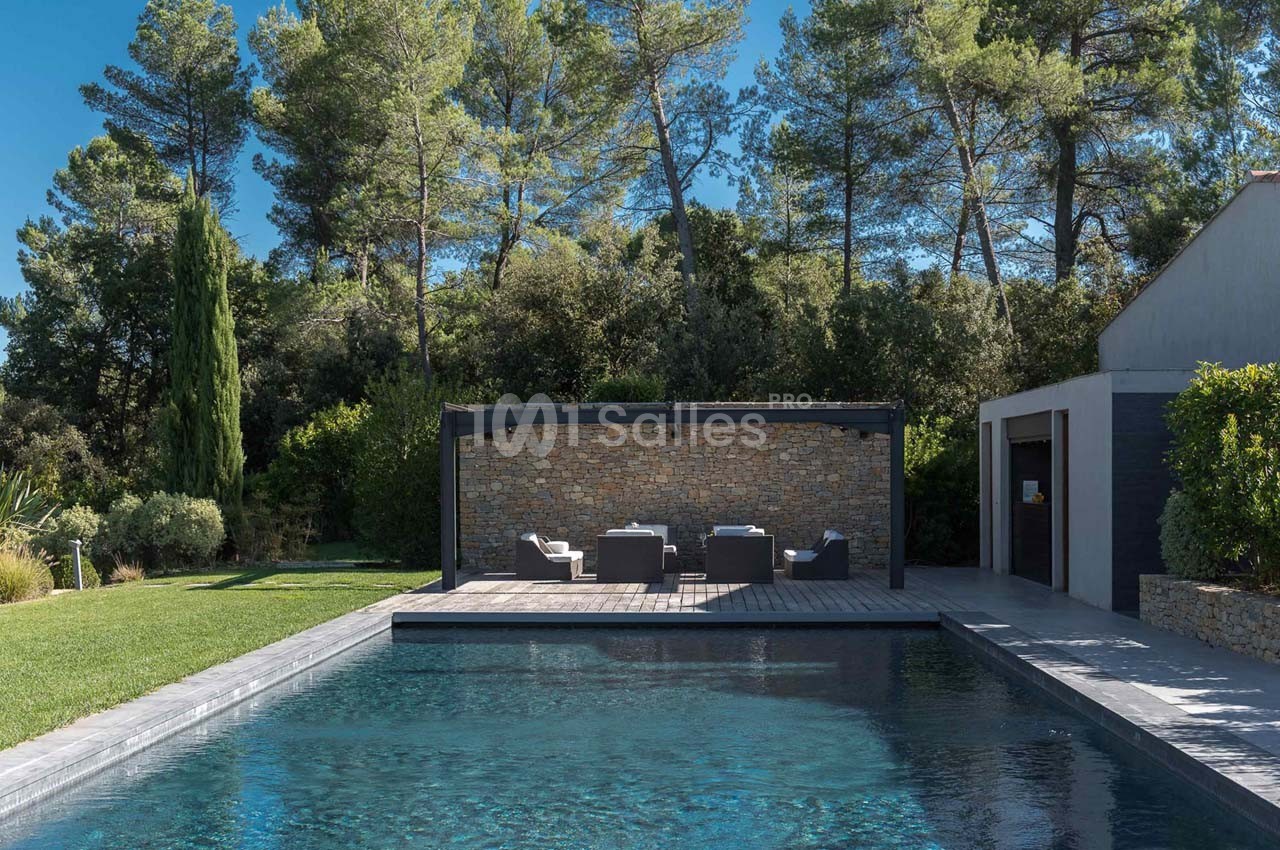 Terrasse avec salon de jardin sous une pergola, piscine au premier plan et arbres en arrière-plan.
