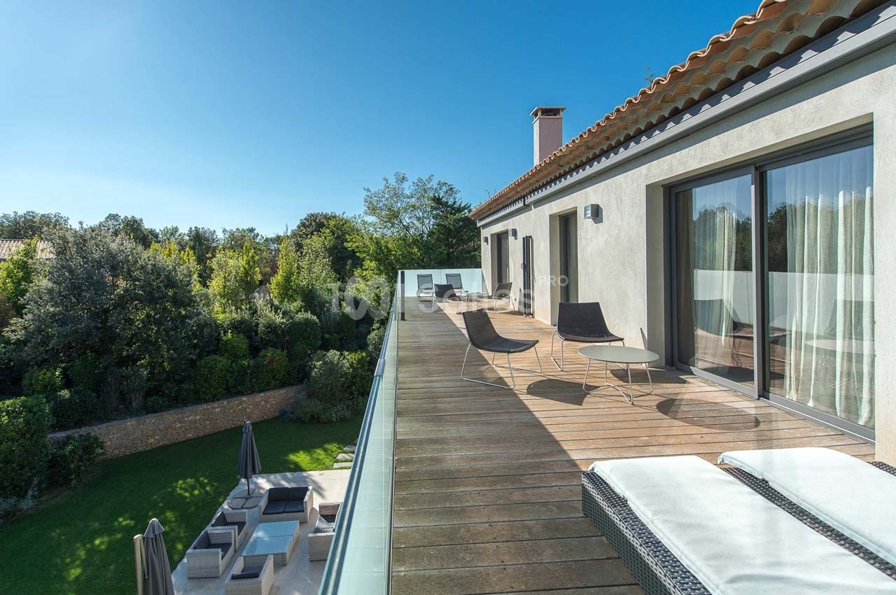 Terrasse en bois avec mobilier extérieur, surplombant un jardin verdoyant par une journée ensoleillée.