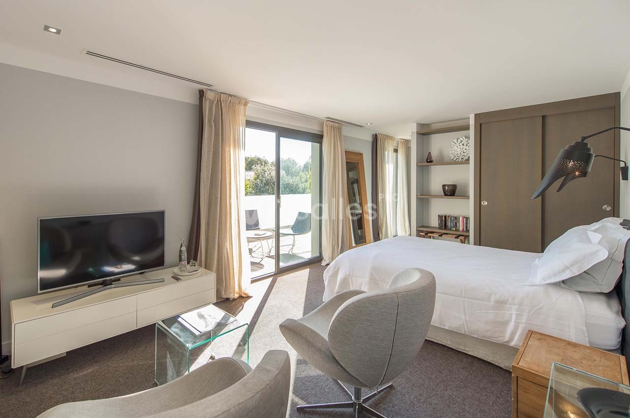 Chambre lumineuse avec lit double, fauteuils, télévision, grande baie vitrée donnant sur une terrasse extérieure.