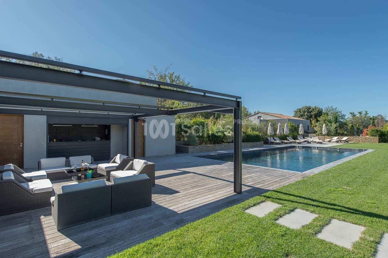 Terrasse avec salon extérieur, pergola, piscine et jardin verdoyant sous un ciel dégagé.