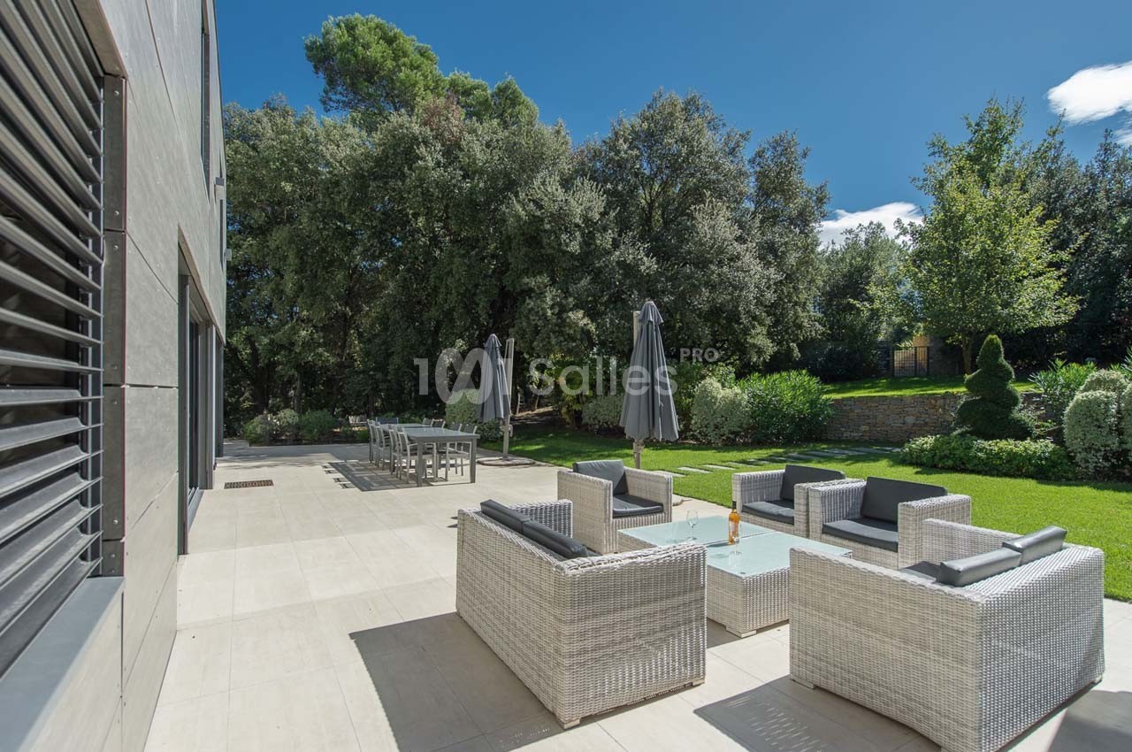 Terrasse aménagée avec mobilier en rotin, parasols et table à manger, entourée d'un jardin arboré sous un ciel bleu.