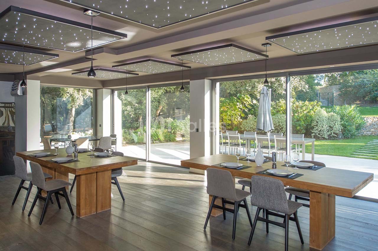 Salle à manger moderne avec tables en bois, chaises grises, grandes baies vitrées et vue sur un jardin verdoyant.