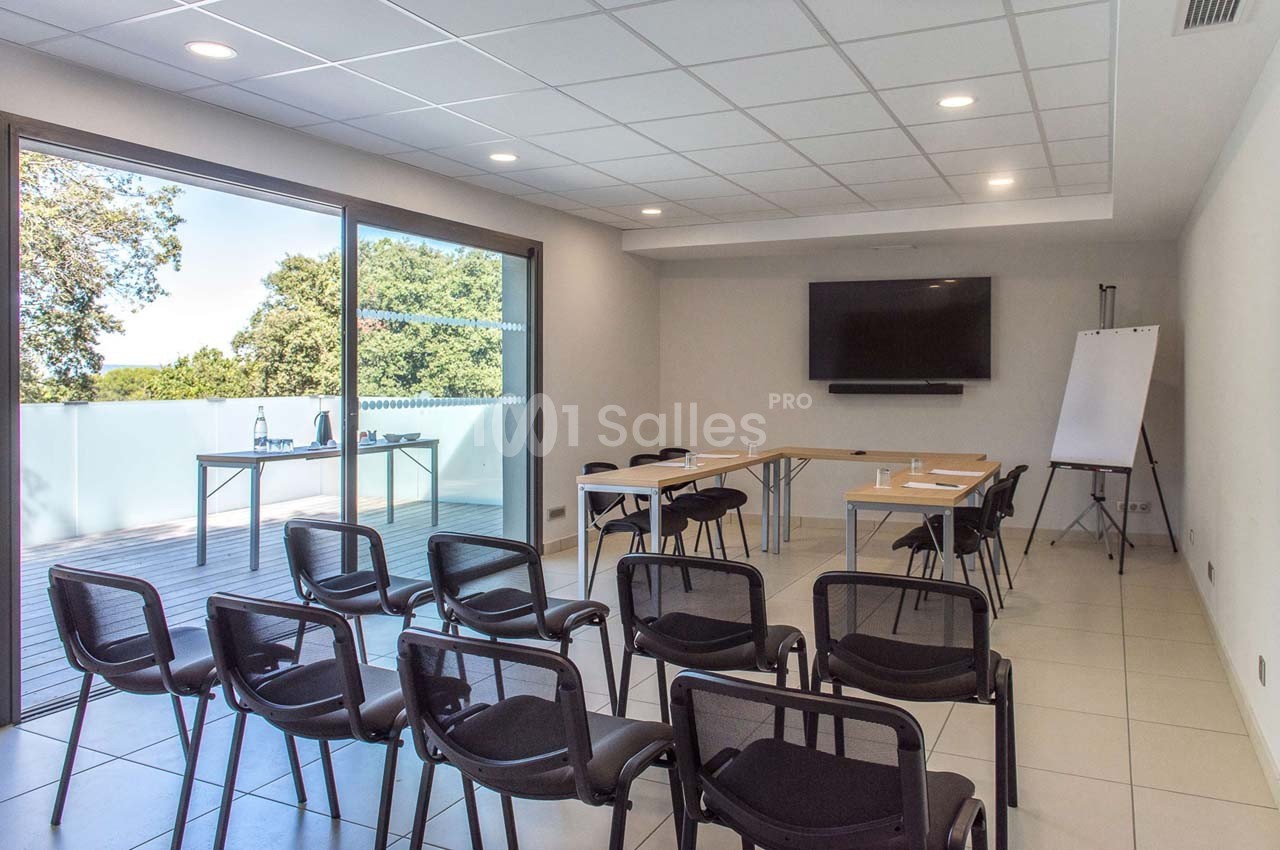 Salle de réunion lumineuse avec chaises alignées, tables disposées en U, tableau blanc et écran mural.