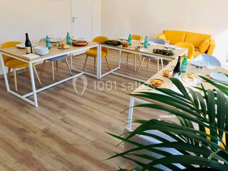 Salle lumineuse avec tables dressées pour un repas, comprenant plats variés, bouteilles et chaises jaunes.