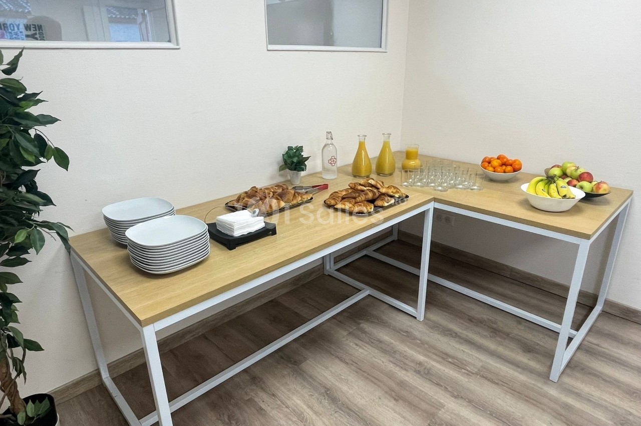 Table avec viennoiseries, assiettes, verres, jus de fruits et corbeilles de fruits dans une salle lumineuse.
