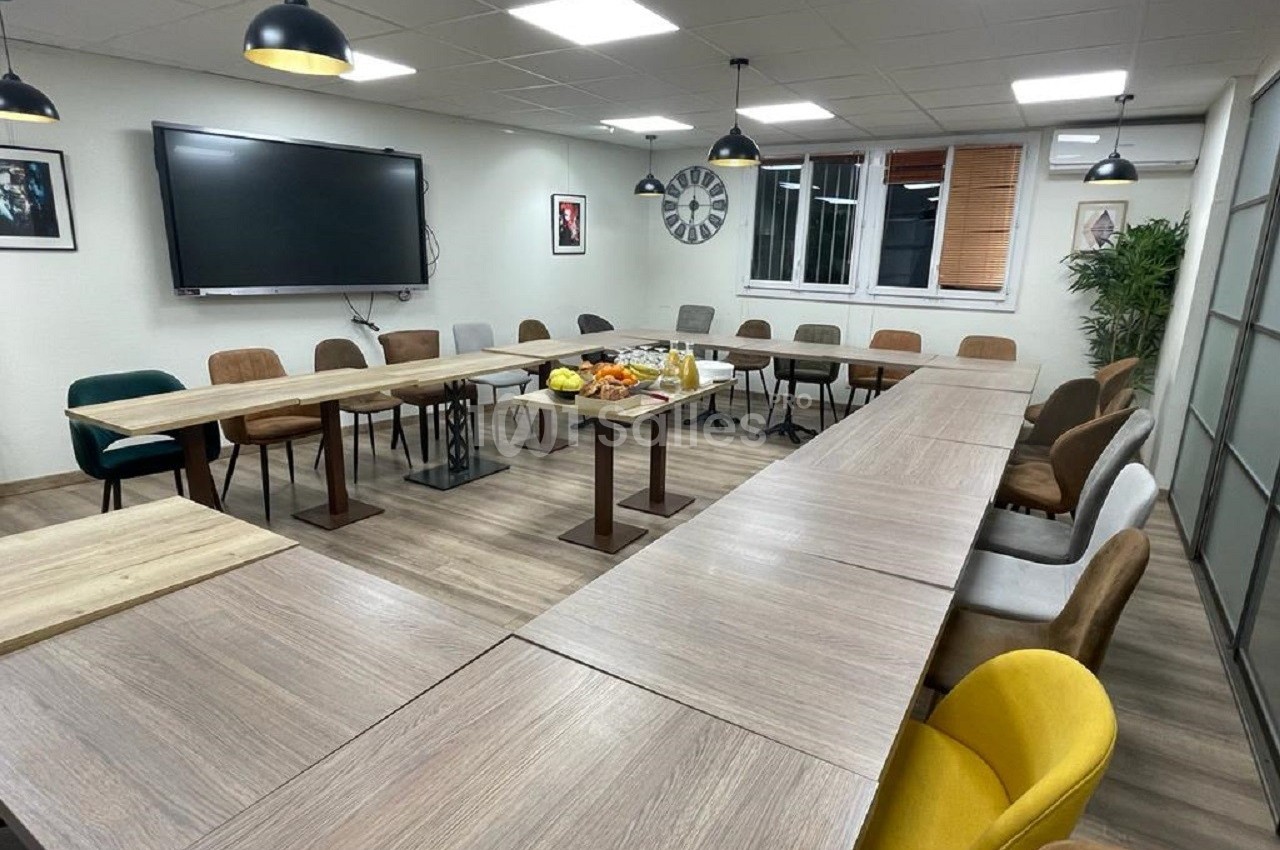 Salle de réunion avec tables disposées en U, chaises colorées, écran mural et plateau central avec collations.