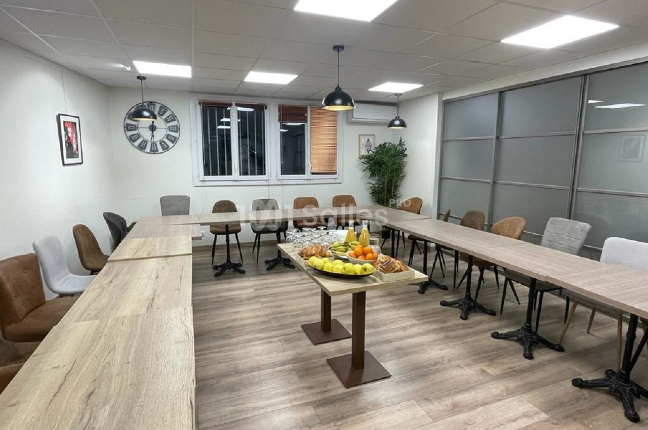 Salle de réunion lumineuse avec tables disposées en U, chaises variées et plateau de fruits au centre.