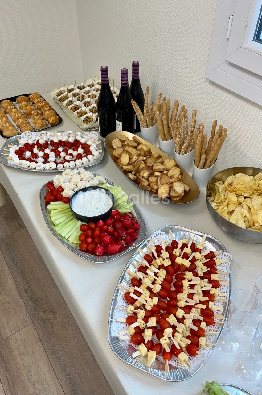 Buffet avec brochettes apéritives, légumes, chips, pains, verrines, et bouteilles de vin rouge sur une table.