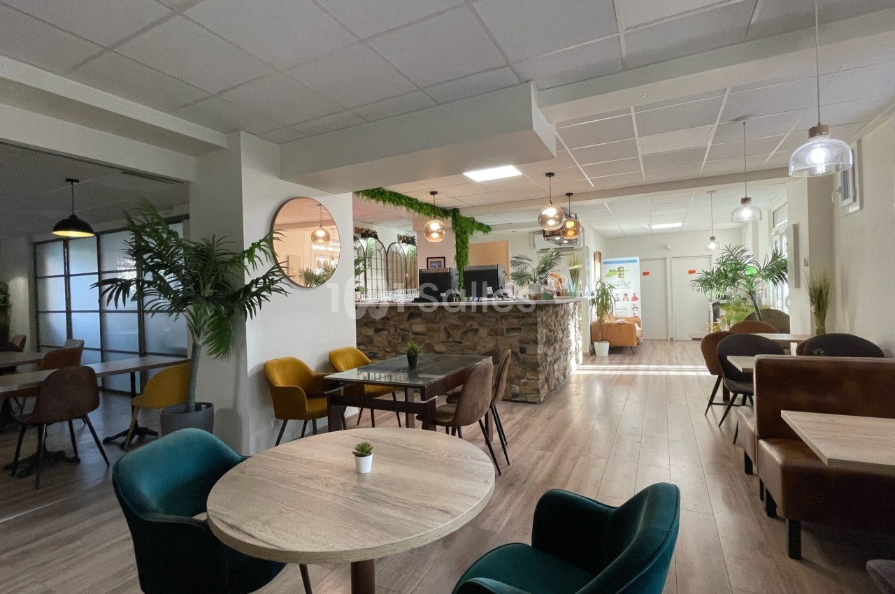 Salle lumineuse avec tables en bois, chaises colorées, plantes décoratives et comptoir en pierre au fond.