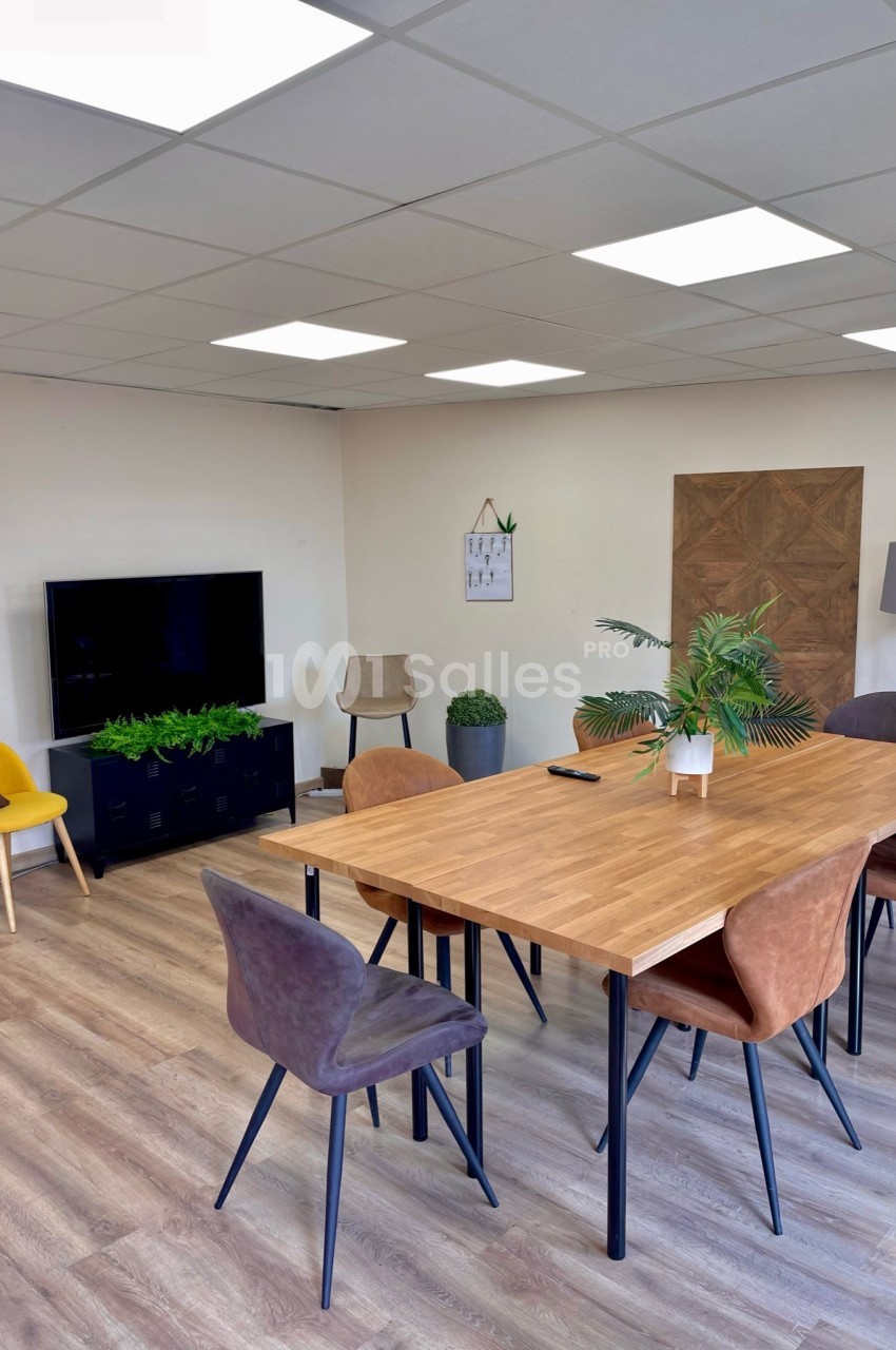 Salle de réunion lumineuse avec table en bois, chaises colorées, plantes décoratives et écran mural.