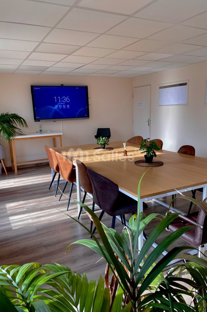 Salle de réunion lumineuse avec une grande table rectangulaire, des chaises, un écran mural et des plantes décoratives.