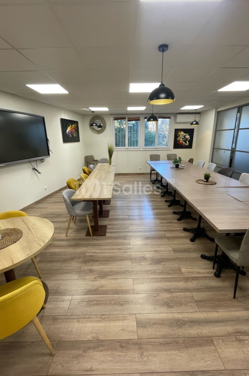 Salle de réunion lumineuse avec tables en bois, chaises colorées, écran mural et grandes fenêtres donnant sur l'extérieur.