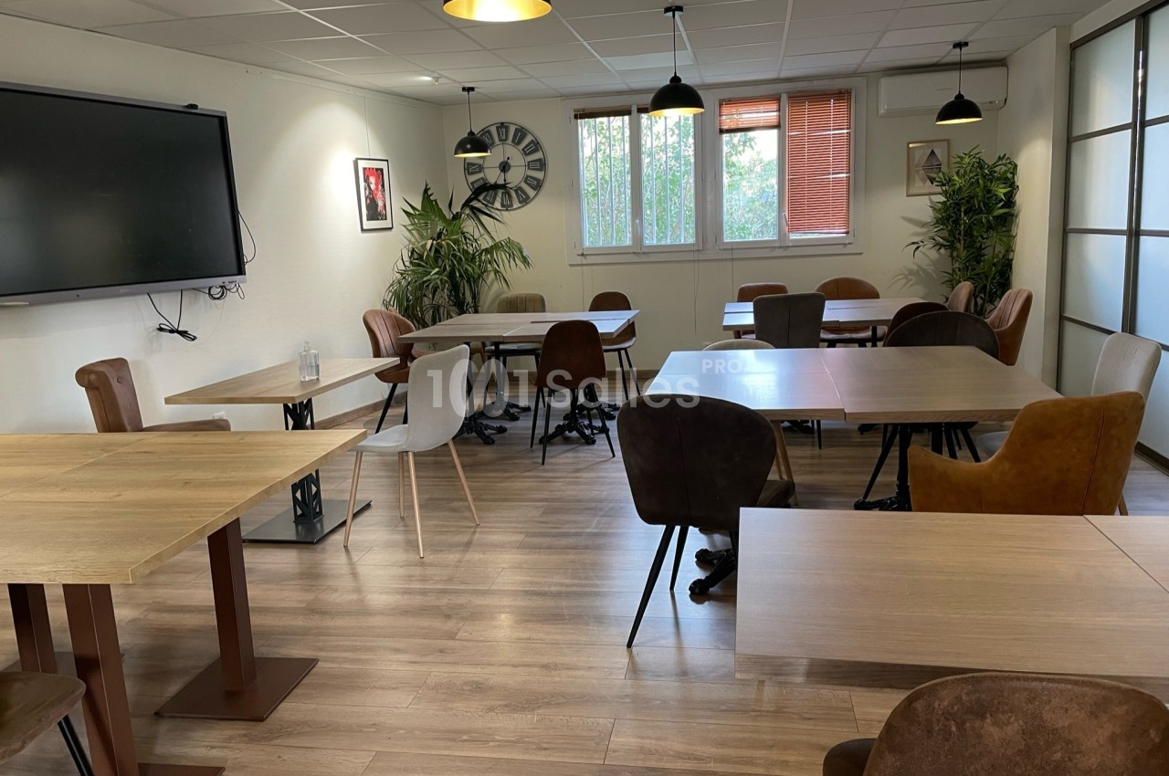 Salle lumineuse avec tables en bois, chaises variées, plantes vertes, tableau mural et grandes fenêtres.