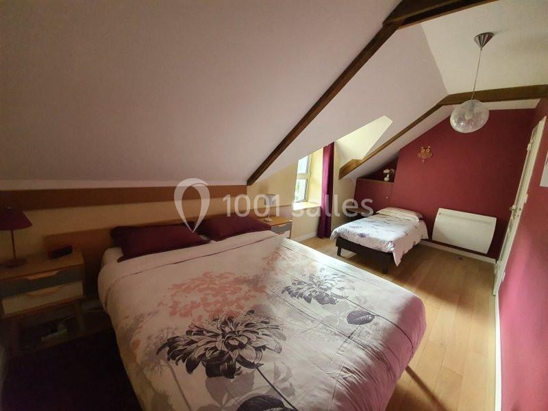 Chambre mansardée avec un lit double, un lit simple, murs rouges et fenêtre laissant entrer la lumière naturelle.