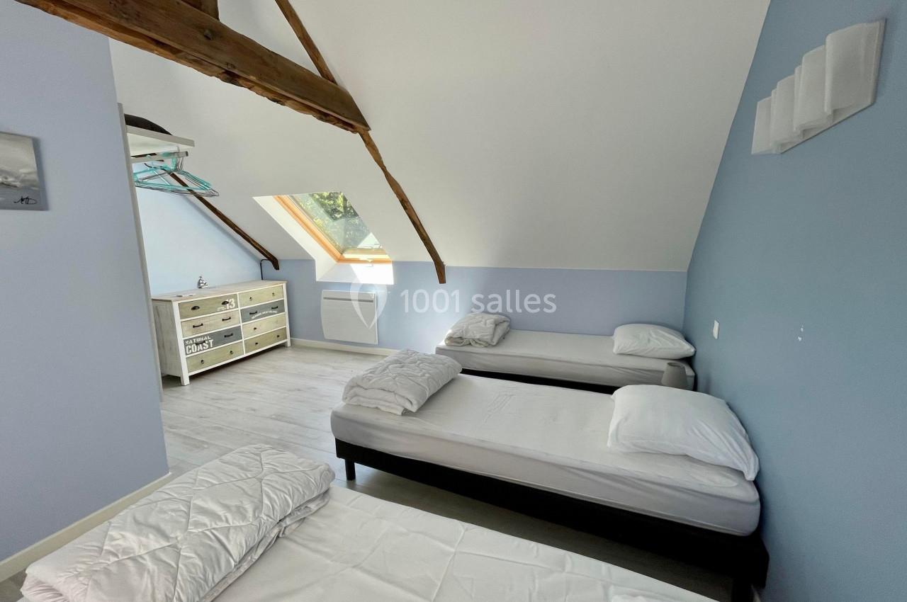 Chambre mansardée avec trois lits simples, murs bleus, poutres apparentes et lumière naturelle provenant d'une fenêtre de…