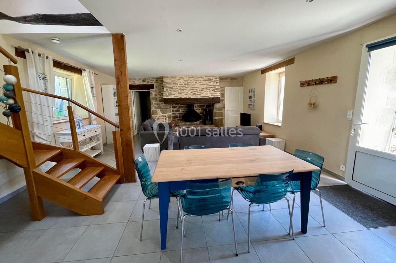 Salle à manger lumineuse avec table en bois, chaises bleues, escalier en bois, et salon avec cheminée en pierre au fond.