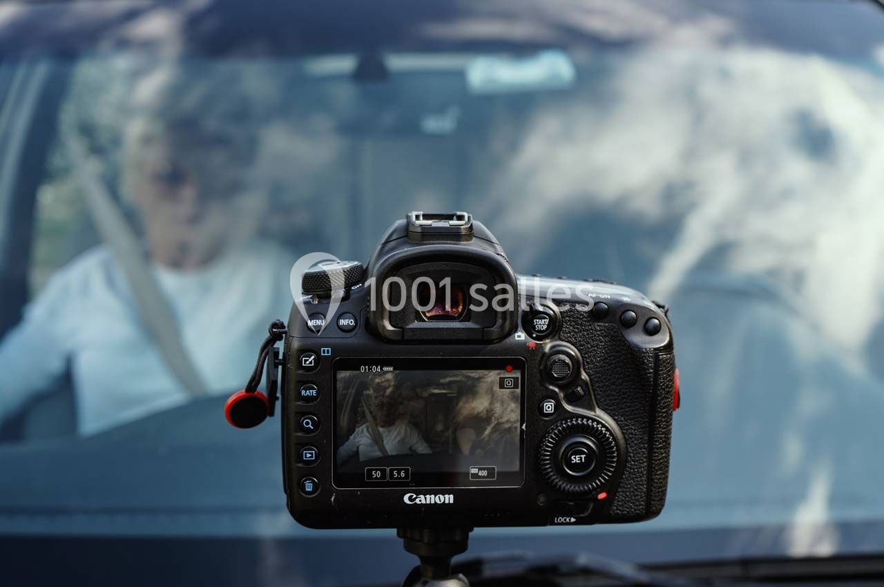 Appareil photo sur trépied cadrant une personne assise dans une voiture, vue à travers le pare-brise.