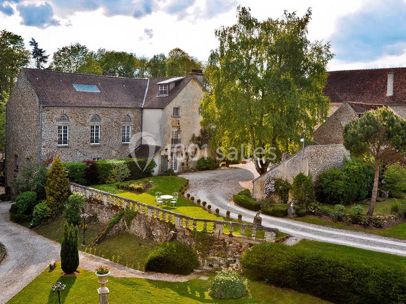 Vue d'un domaine rural avec bâtiments en pierre, jardin aménagé, allées gravillonnées et arbres environnants.