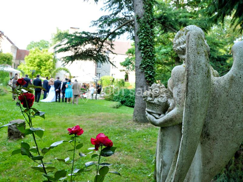 Statue d'ange tenant des fleurs dans un jardin, avec un mariage en arrière-plan sous des arbres.