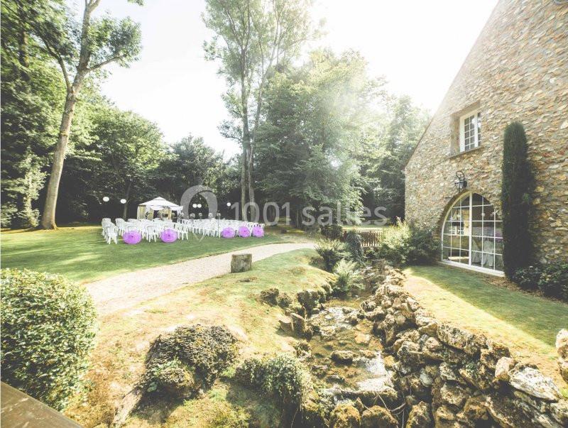 Jardin avec pelouse, chaises blanches disposées pour un événement, bâtiment en pierre et ruisseau au premier plan.