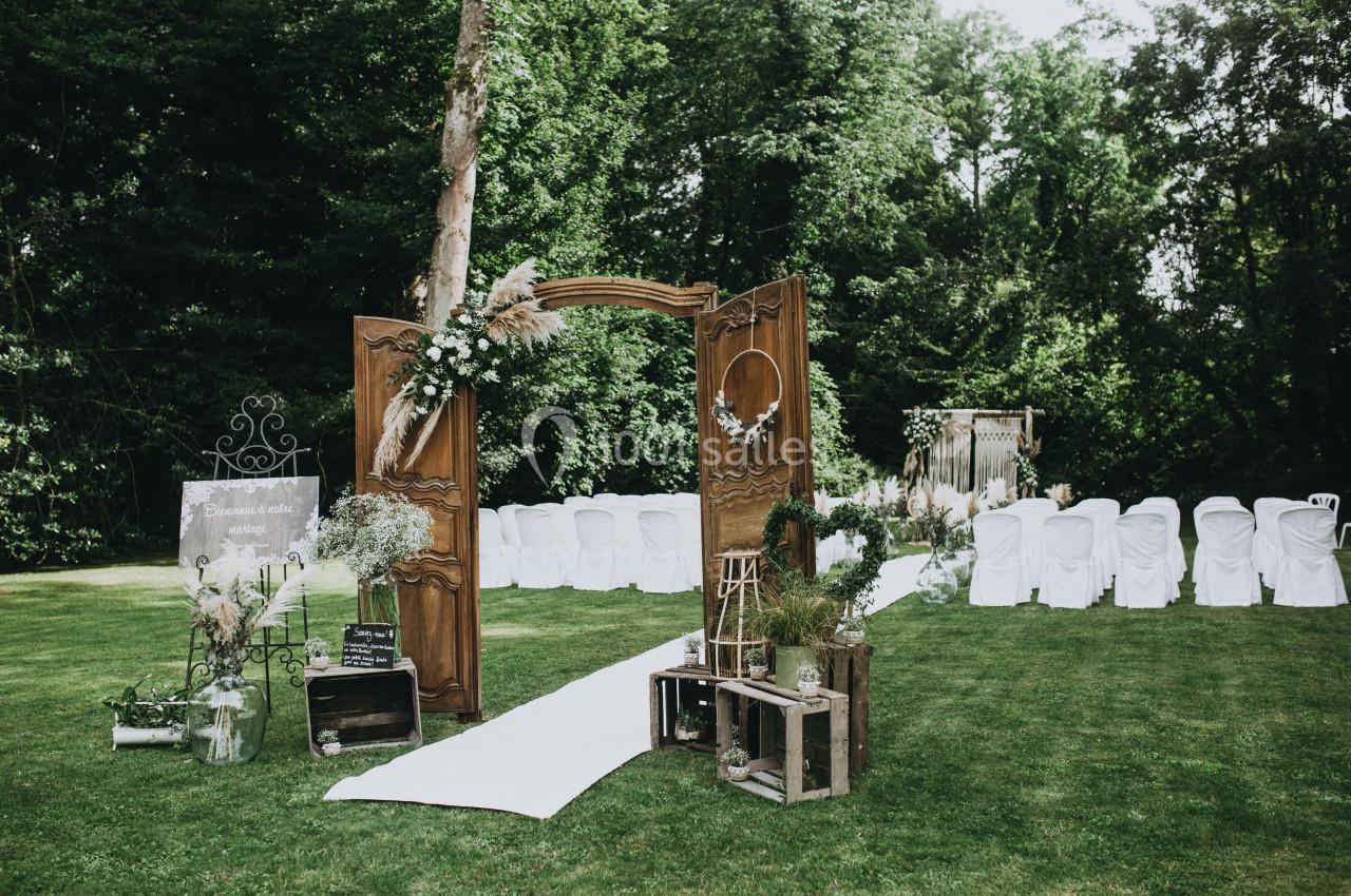 Allée de cérémonie en plein air avec chaises blanches, arche en bois décorée de fleurs et verdure environnante.