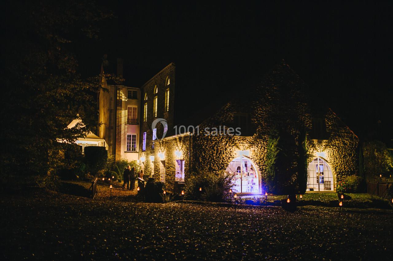 Bâtiments en pierre éclairés la nuit, avec des fenêtres illuminées et un jardin sombre au premier plan.