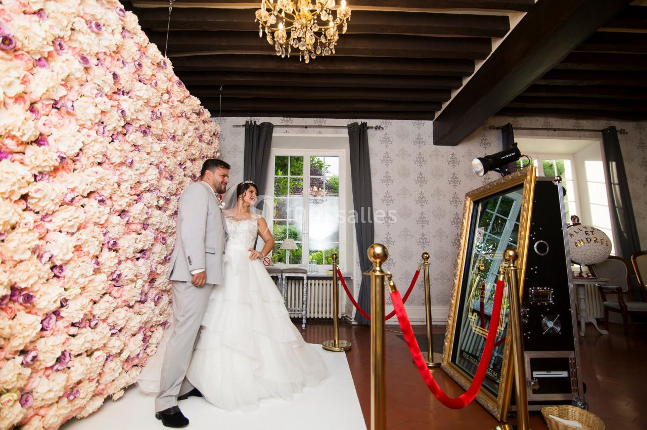 Un couple pose devant un mur de fleurs et un miroir interactif dans une salle décorée avec un lustre et des poutres…