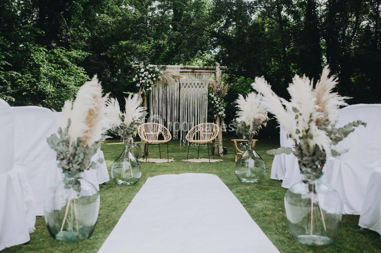 Allée centrale menant à une arche décorée de fleurs et de macramé, entourée de chaises blanches et de vases avec des herbes…