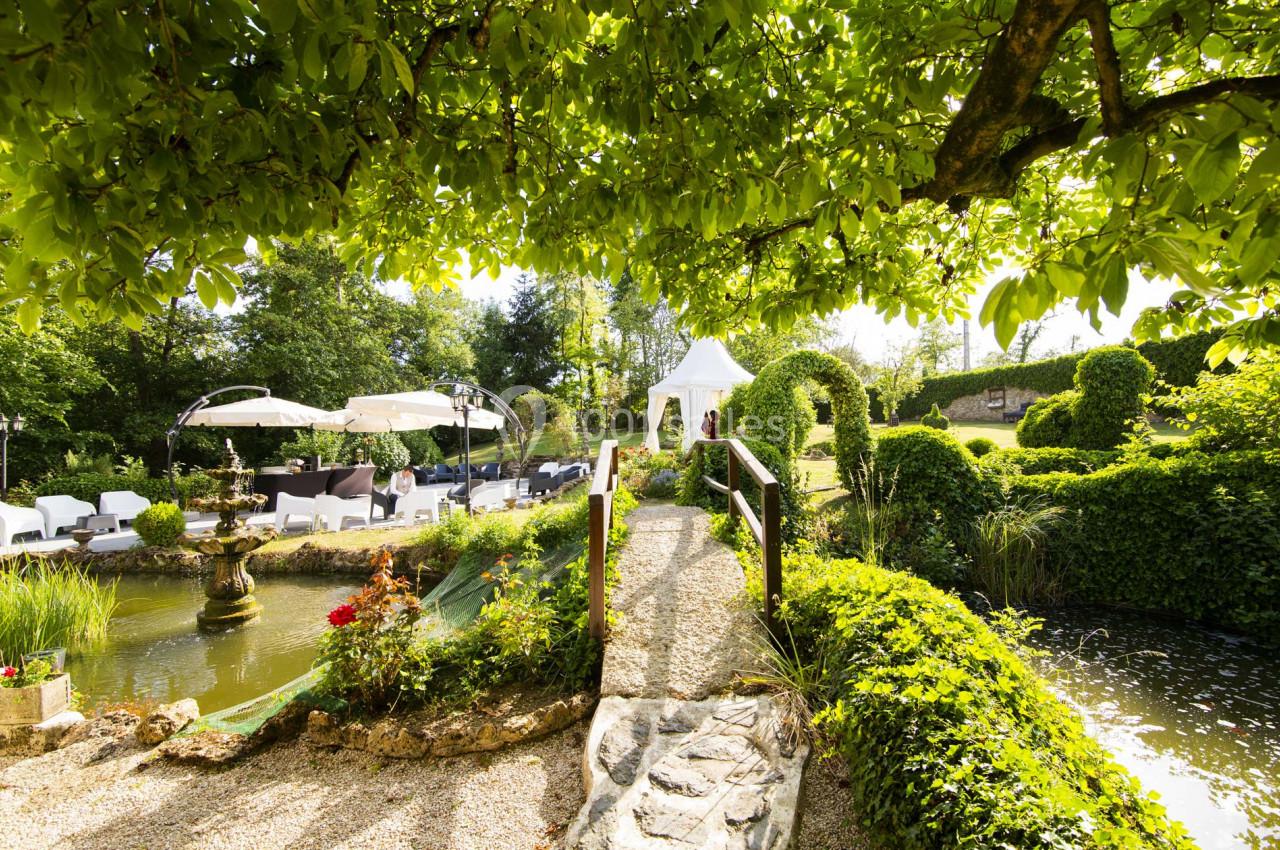 Jardin verdoyant avec un pont en bois, une fontaine, des parasols et un pavillon blanc sous un ciel ensoleillé.