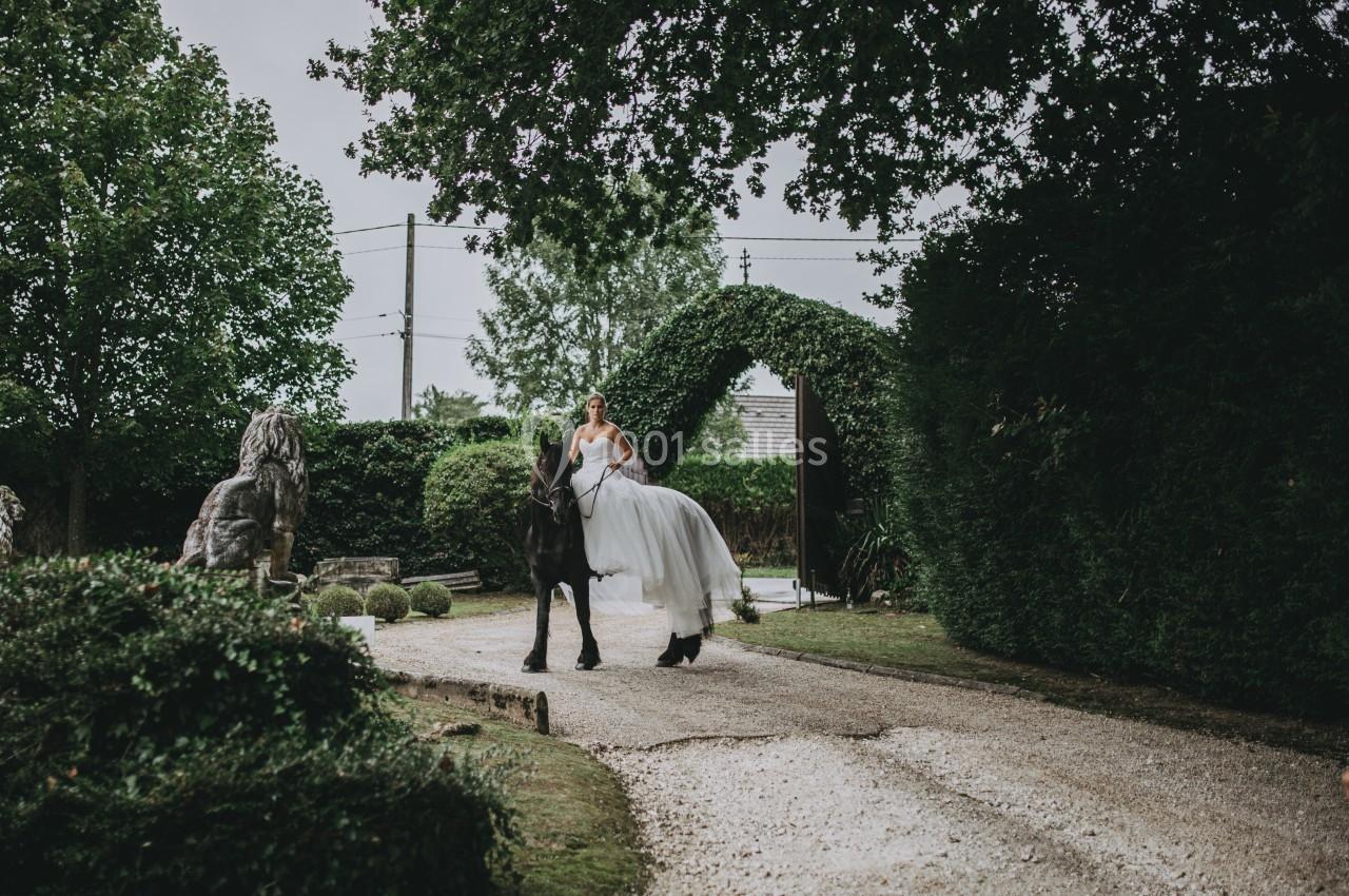 Une mariée en robe blanche monte un cheval noir sur une allée bordée de verdure, sous un ciel nuageux.