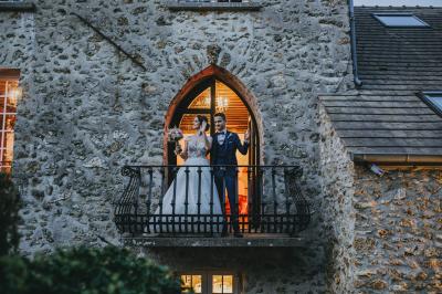 Un couple en tenue de mariage se tient sur un balcon en pierre devant une fenêtre illuminée.