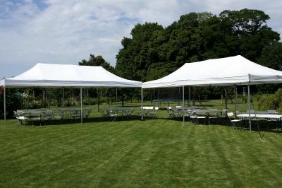 Deux tentes blanches montées sur une pelouse avec des tables et des chaises disposées autour, entourées de verdure.