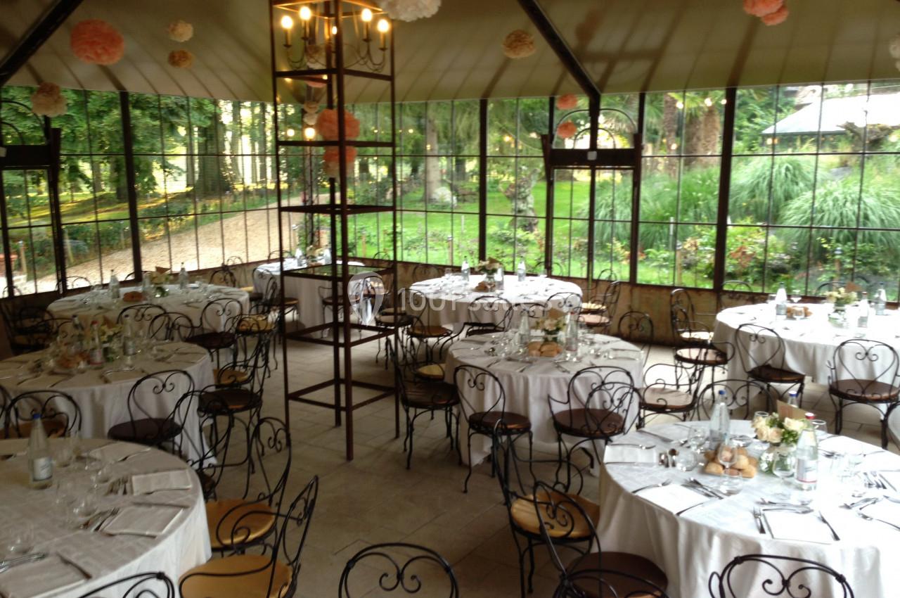 Salle de réception lumineuse avec tables rondes dressées, chaises en fer forgé et vue sur un jardin verdoyant.