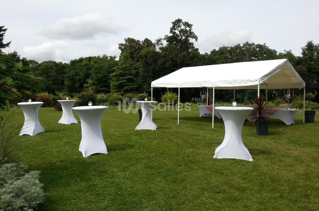 Tente blanche et tables hautes couvertes de nappes blanches disposées sur une pelouse dans un jardin verdoyant.