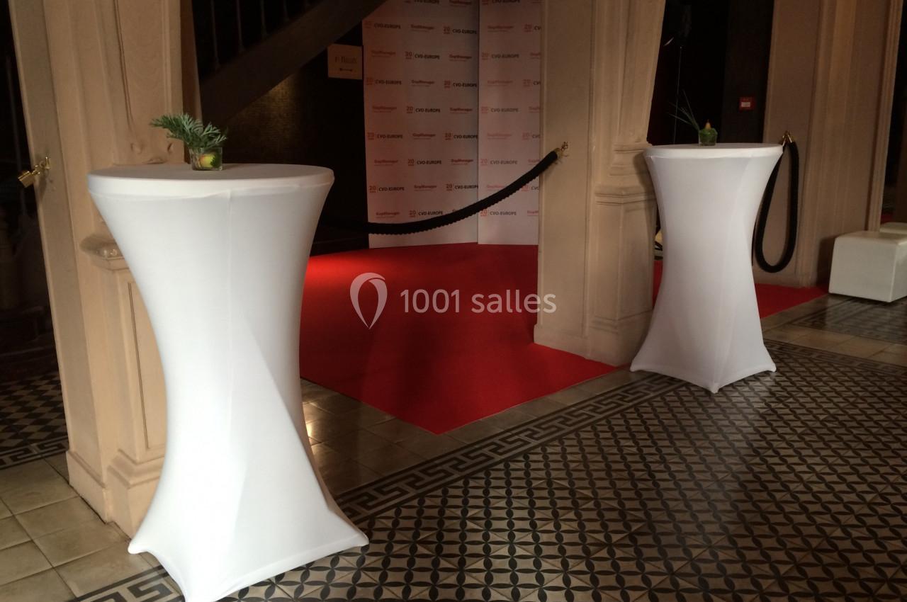 Deux tables hautes drapées de blanc encadrent un tapis rouge dans un espace intérieur au sol carrelé.